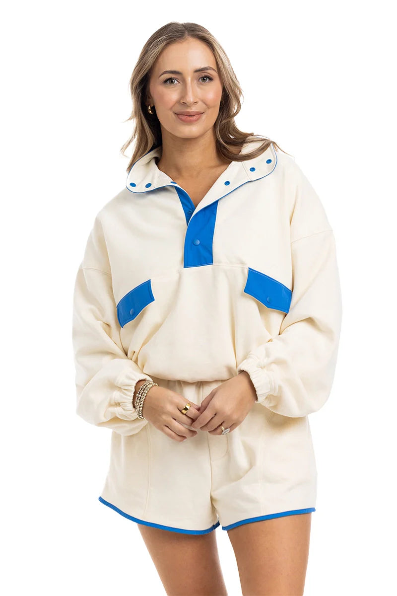 A woman in Katydids Yellow  Blue Quarter Snap Athletic Set—featuring a cream long-sleeve pullover with blue details and matching shorts—smiles at the camera, hands clasped.
