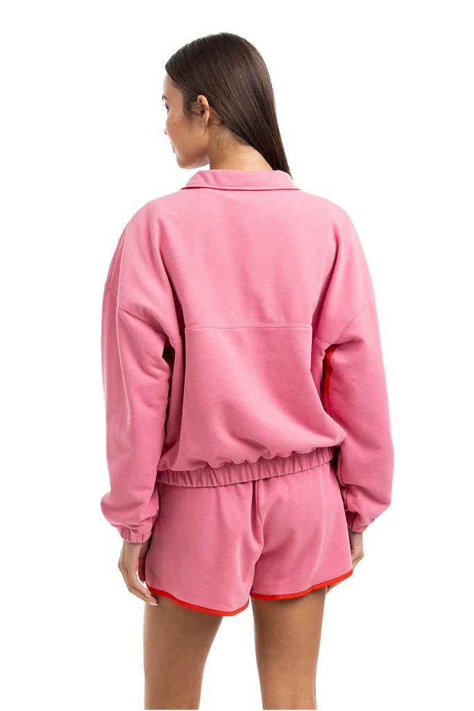 A woman with straight brown hair models the Katydid Pink  Orange Quarter Snap 2 Piece Athletic Set—vibrant loungewear in ultra-soft performance fabric. She faces away from the camera, her left hand resting at her side.