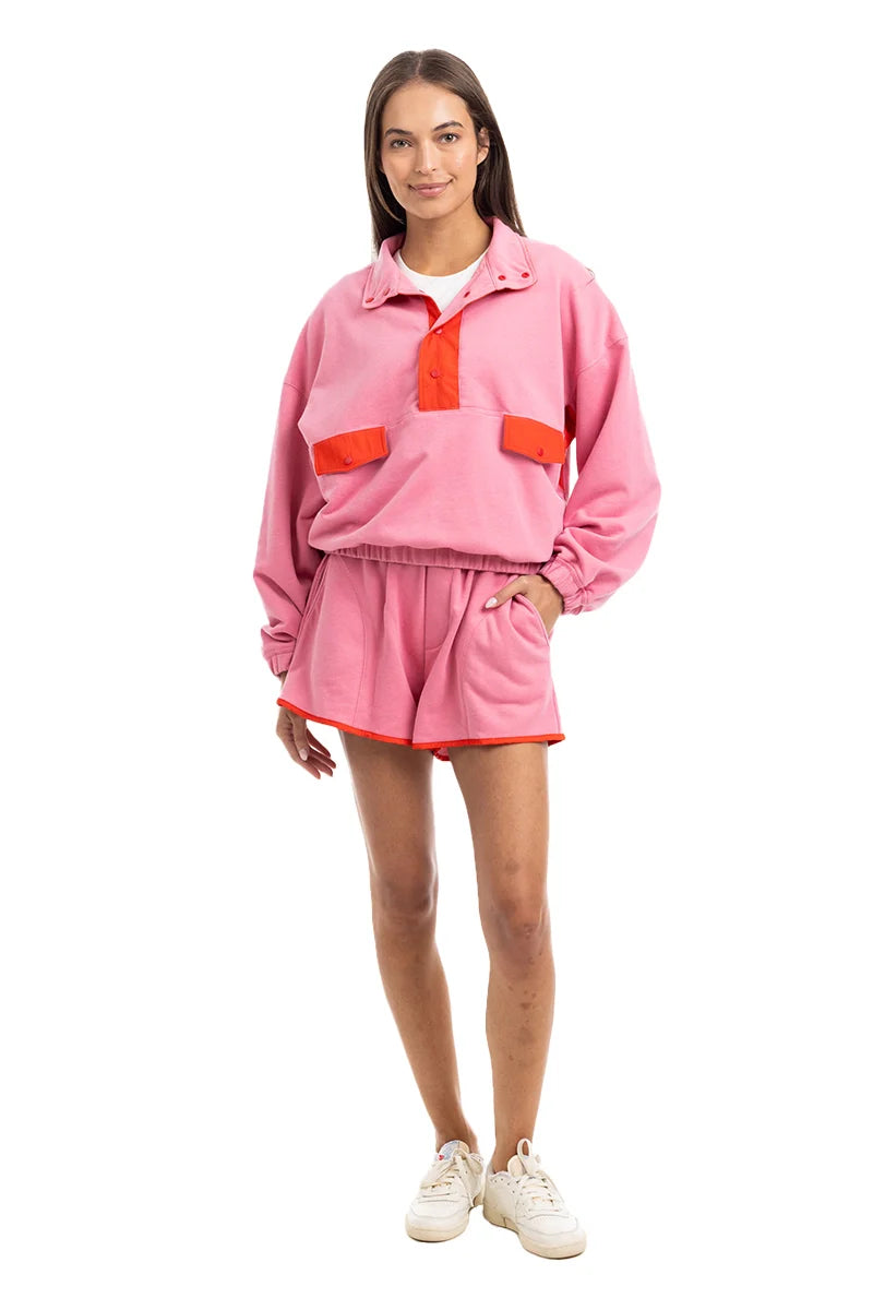 A woman with straight brown hair and white sneakers stands facing forward in the Katydid Pink  Orange Quarter Snap 2 Piece Athletic Set—vibrant, ultra-soft performance loungewear—against a plain white background.
