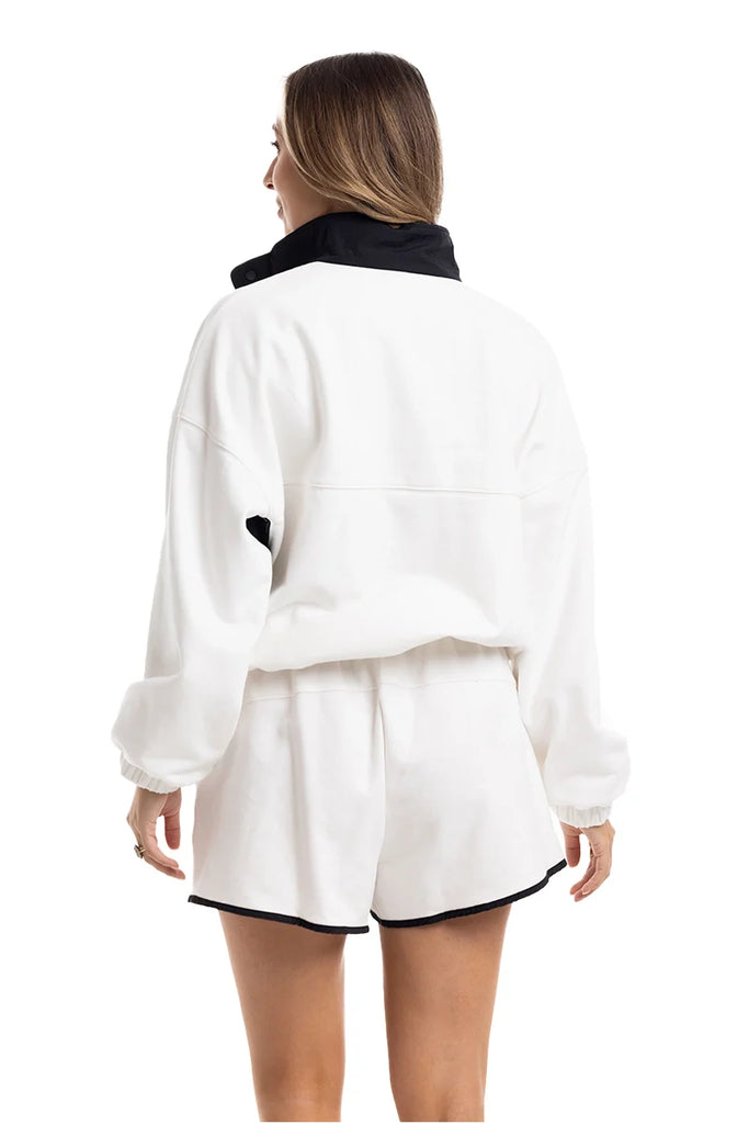 A woman, her light brown hair down, stands with her back to the camera in Katydid’s White  Black Quarter Snap Set—an athletic fit featuring a loose white pullover with black collar and matching white shorts with black trim.