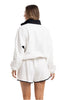 A woman, her light brown hair down, stands with her back to the camera in Katydid’s White  Black Quarter Snap Set—an athletic fit featuring a loose white pullover with black collar and matching white shorts with black trim.