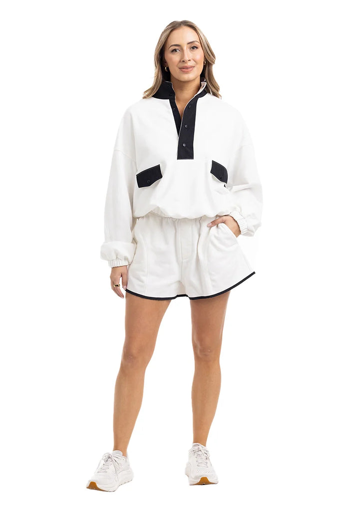 A woman smiles in the Katydid White  Black Quarter Snap Set— a sporty lounge outfit with a white long-sleeve pullover with black accents, matching white shorts with black trim, and white sneakers. She poses against a white background.