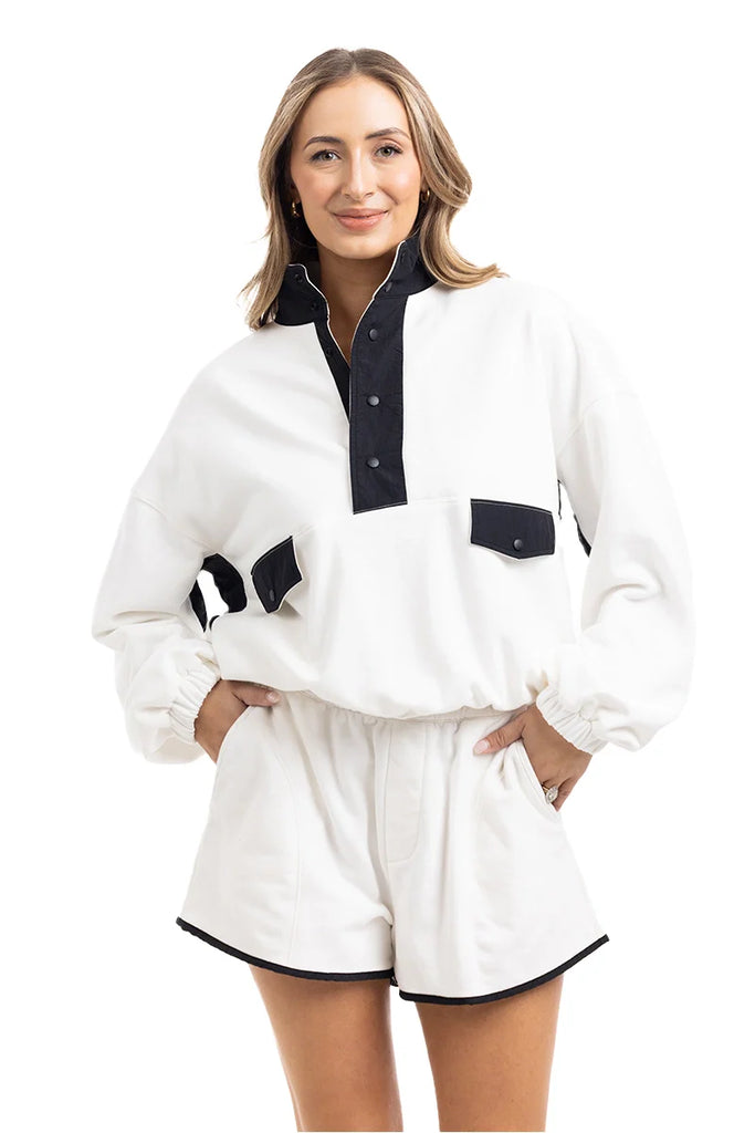A woman smiles in the Katydid White  Black Quarter Snap Set: a sporty long-sleeve pullover with black buttons and trim, paired with high-waisted shorts edged in black. She stands against a plain white background.