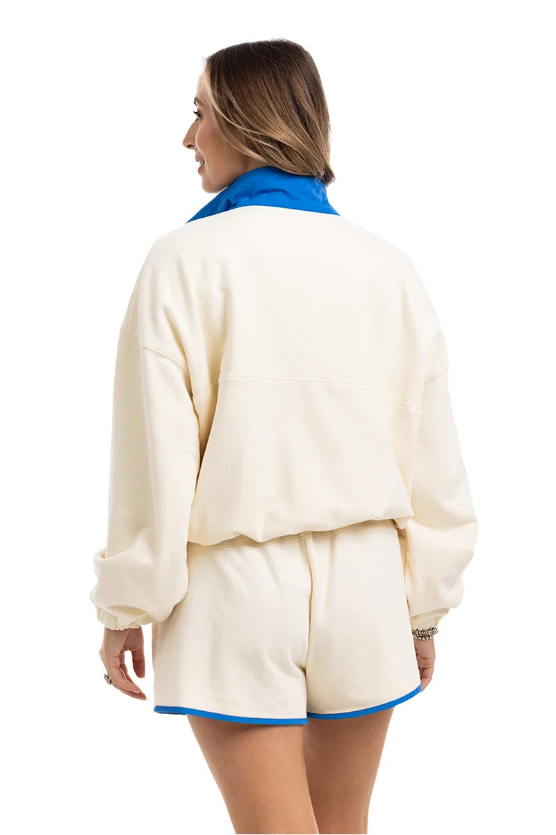 A woman with light brown hair, accessorized with bracelets, models the Katydid Yellow  Blue Quarter Snap Athletic Set—featuring a matching long-sleeve top and shorts with blue trim and collar—in an athletic fit.