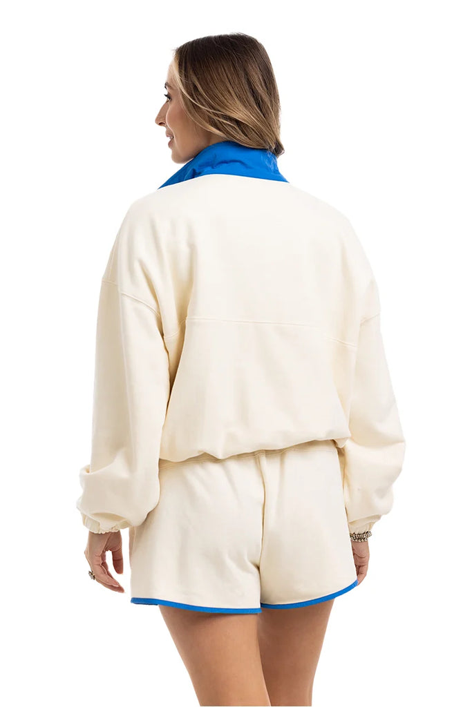 A woman with light brown hair, accessorized with bracelets, models the Katydid Yellow  Blue Quarter Snap Athletic Set—featuring a matching long-sleeve top and shorts with blue trim and collar—in an athletic fit.