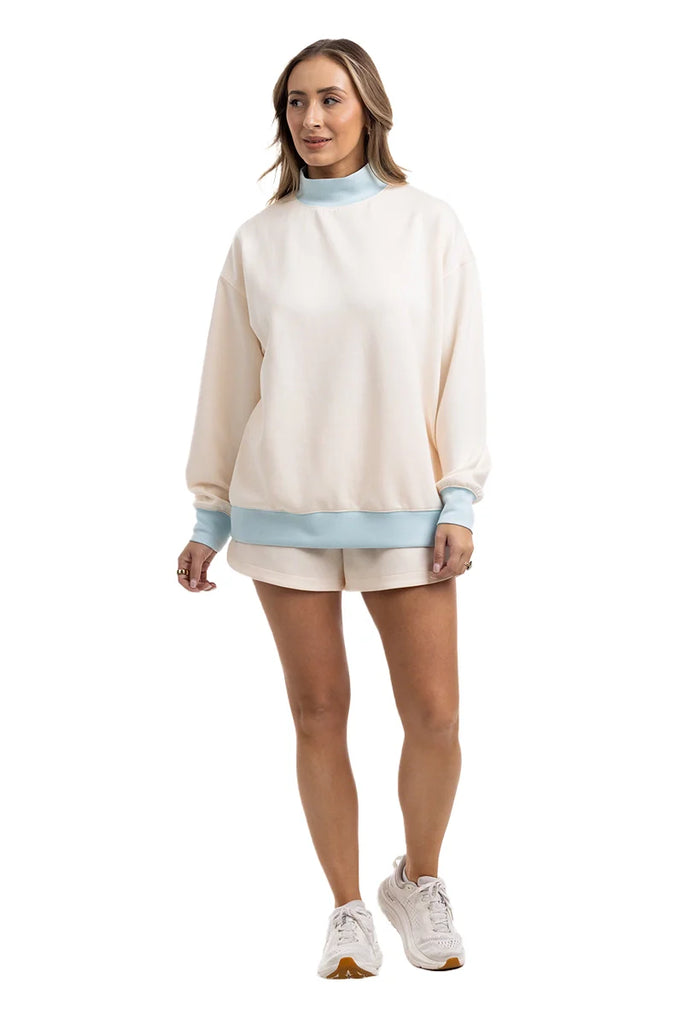 A woman models the Katydid Beige  Light Blue Duo Mockneck Matching Set—featuring a cream oversized top with blue cuffs and hem, cream shorts, and white sneakers—against a white background.