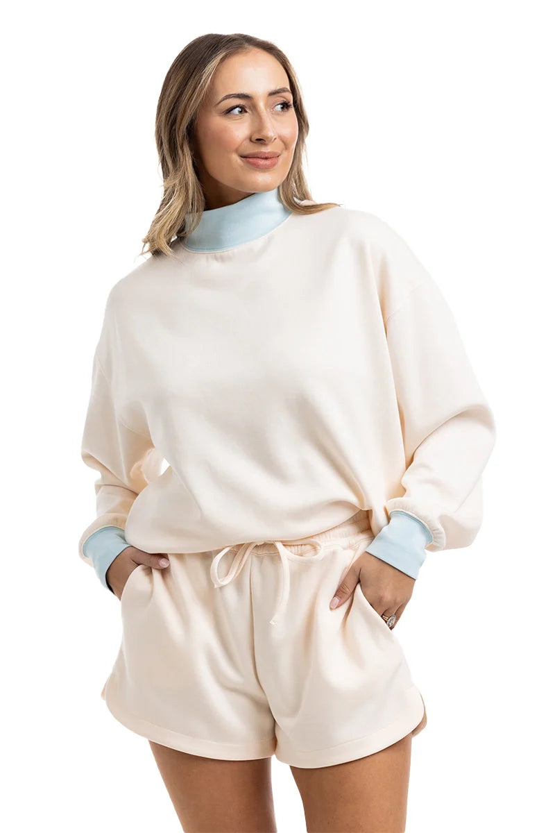 A woman wears the Katydid Beige  Light Blue Duo Mockneck Matching Set, featuring a mockneck sweatshirt and cream shorts. She stands with a hand in her pocket, smiling and looking to the side against a white background.
