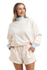 A woman wears the Katydid Beige  Light Blue Duo Mockneck Matching Set, featuring a mockneck sweatshirt and cream shorts. She stands with a hand in her pocket, smiling and looking to the side against a white background.