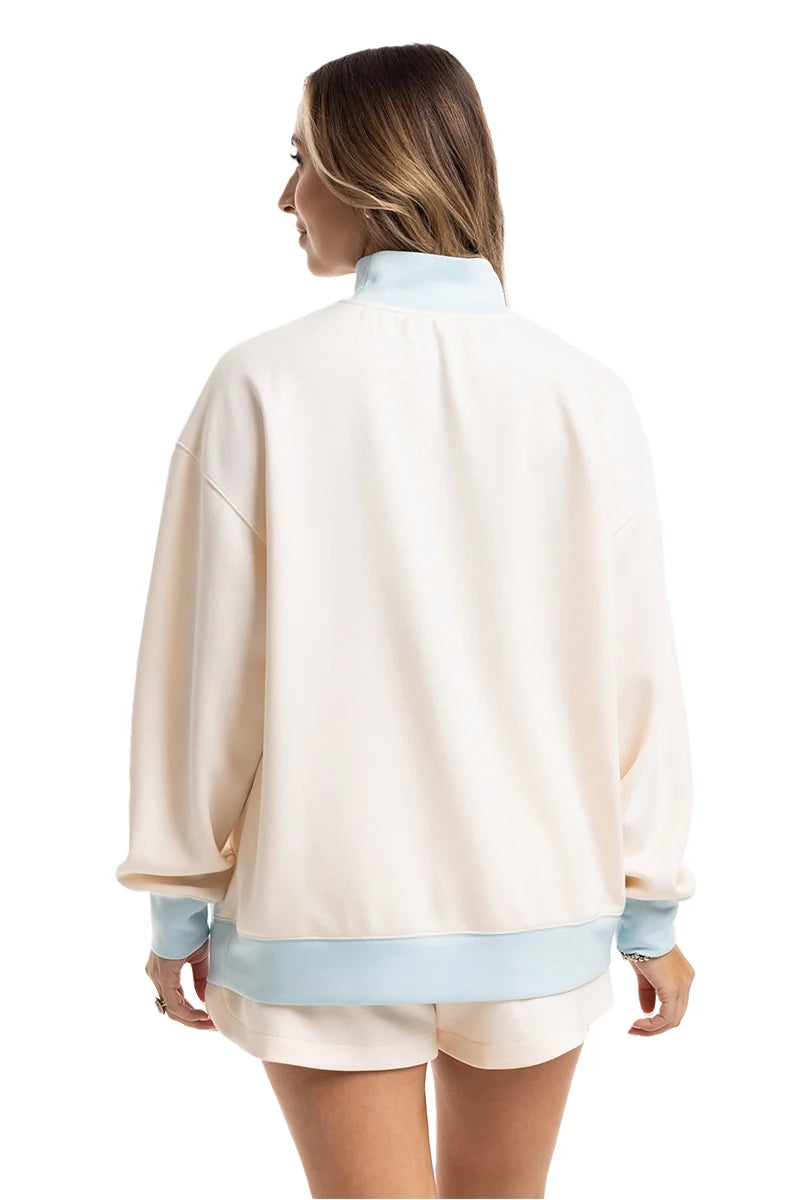 A woman stands facing away, wearing the Katydid Beige  Light Blue Duo Mockneck Matching Set featuring a loose mockneck top with cream shorts and light blue cuff and hem accents.