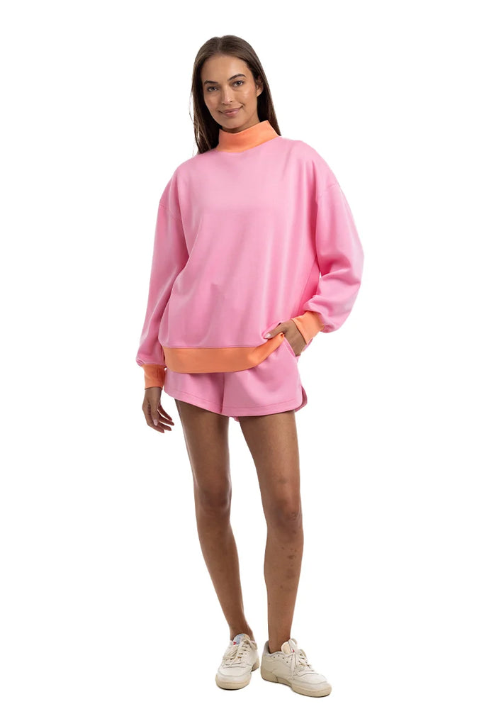 A person models the Katydid Pink  Orange Duo Mockneck Sweatset—featuring a loose sweatshirt with orange cuffs and collar, matching pink shorts with an orange waistband—and white sneakers, all set against a white background.