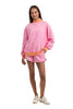 A person models the Katydid Pink  Orange Duo Mockneck Sweatset—featuring a loose sweatshirt with orange cuffs and collar, matching pink shorts with an orange waistband—and white sneakers, all set against a white background.