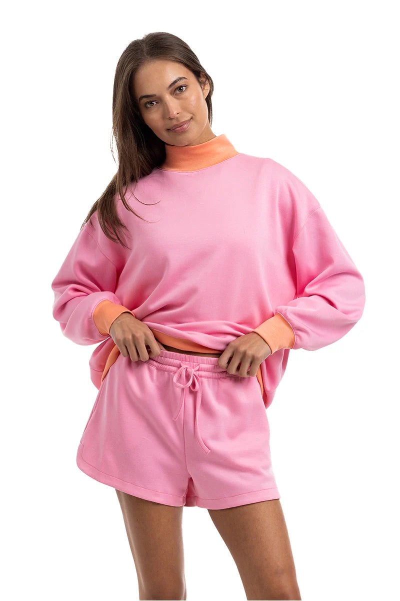 A woman smiles against a white background, wearing the Katydid Pink  Orange Duo Mockneck Sweatset—an easygoing loungewear combo featuring a relaxed mockneck sweatshirt and drawstring shorts—while holding the hem of her top.