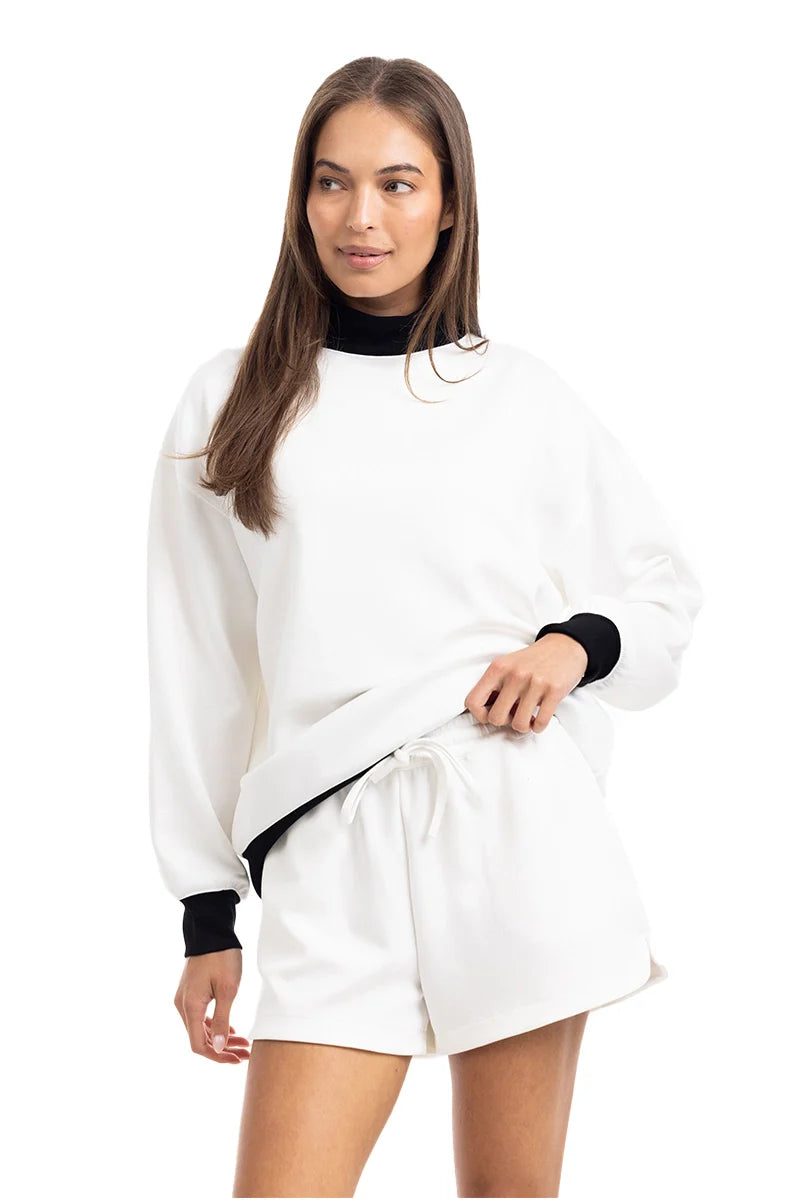 A woman models the Katydid White  Black Duo Mockneck Set—featuring a white sweatshirt with black cuffs and collar and matching shorts—standing against a plain white background, gazing to the side with a slight smile.