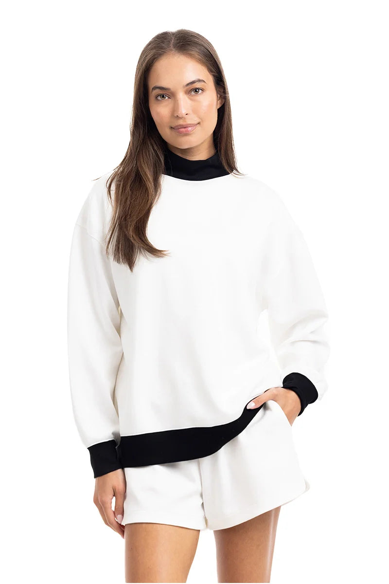 A woman in the Katydid White  Black Duo Mockneck Set—featuring a mockneck top with black collar and cuffs and matching white shorts—stands smiling with one hand in her pocket against a plain white background.