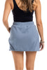 A woman with long, wavy hair models the Katydid Steel Blue Mini Bubble Skirt, paired with a sleeveless black top. Posed facing away, she showcases the skirt’s playful style and bracelets on her right wrist.