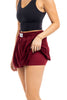 A person in a Katydid Wine Bubble Skirt Skort lifts the top layer to show matching built-in compression shorts. The crop top is black, and the background is white.