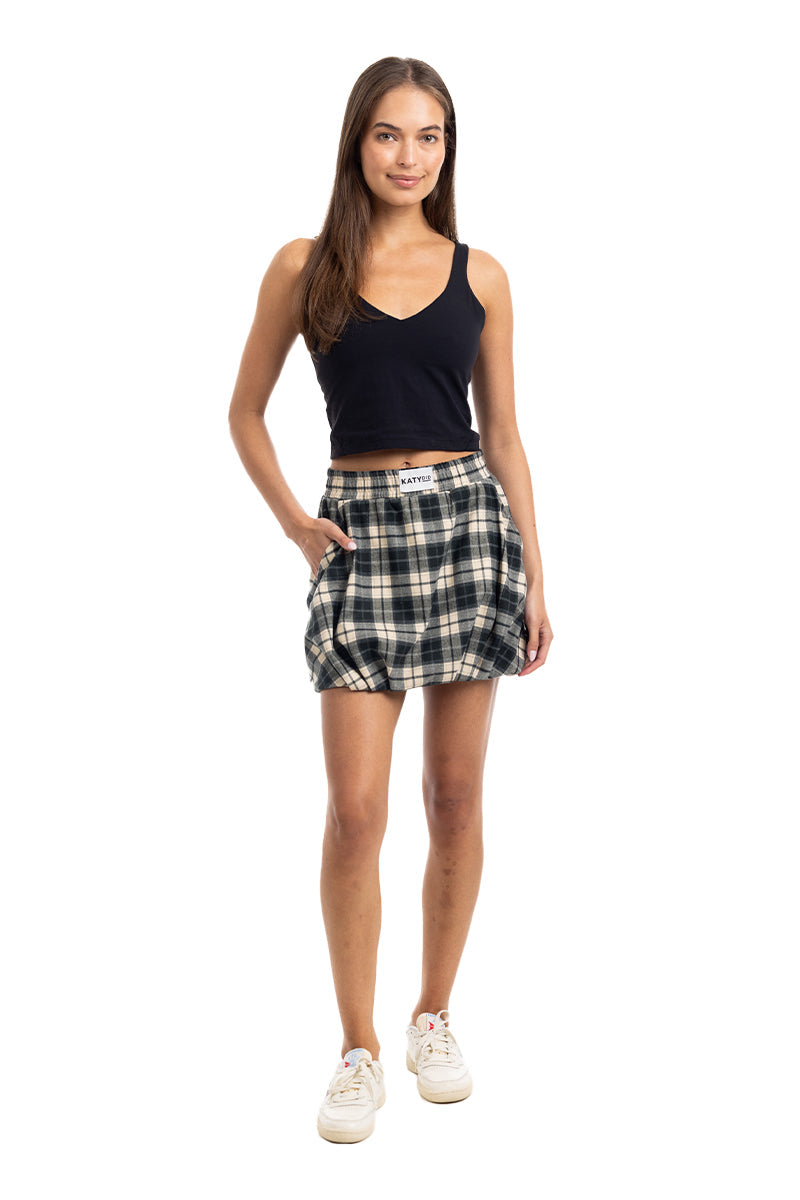 A woman with long brown hair stands facing forward, smiling slightly. She wears a Katydid Black Autumn Plaid Skirt with Shorts in green and white, a black sleeveless V-neck top, and white sneakers against a white background.