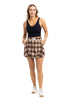 A woman smiles against a white background, wearing the Katydid Brown Plaid Bubble Hem Skirt with pockets, a sleeveless black top, and white sneakers. One hand is in her pocket, showcasing this stylish autumn-ready look.