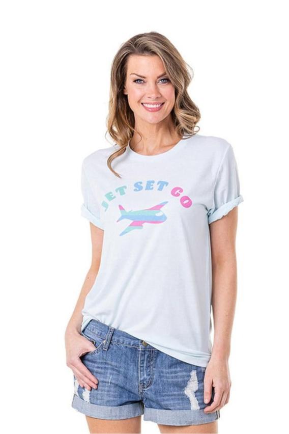 A smiling woman with wavy light brown hair models the Katydid Jet Set Go Women’s T-Shirt, featuring a light blue hue, round neck, and airplane graphic, paired with denim shorts against a white background.