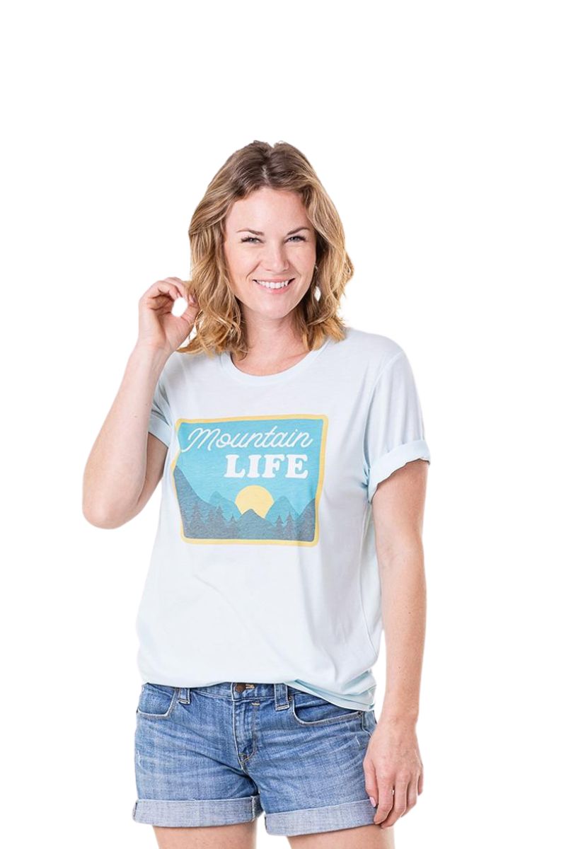 A smiling woman in shoulder-length hair models the Katydid Mountain Life Graphic T-Shirt in light blue with a mountain design, unisex fit, and denim shorts, posed against a white background.