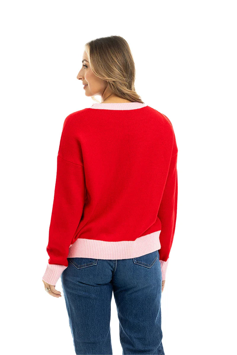 A woman with long light brown hair models the Katydid Merry Embroidered Sweater Christmas Woman, featuring pink cuffs and waistband, paired with blue jeans. She stands facing away and looks over her left shoulder against a white background.