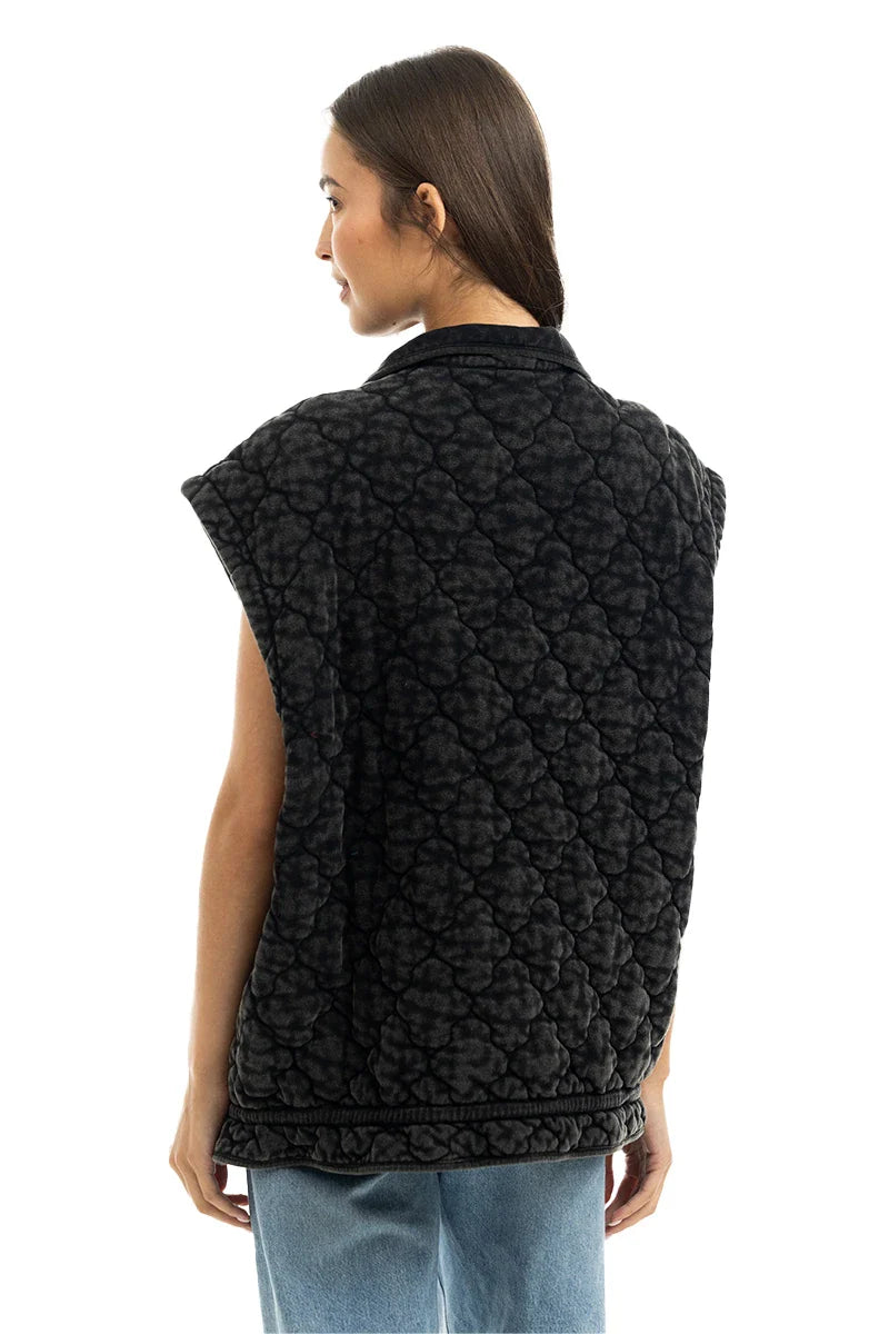A woman with straight brown hair faces away, wearing a Katydid Charcoal Oversized Garment Washed Quilted Vest with wide shoulders over light blue jeans. The vest features a black cloud pattern. The background is white.