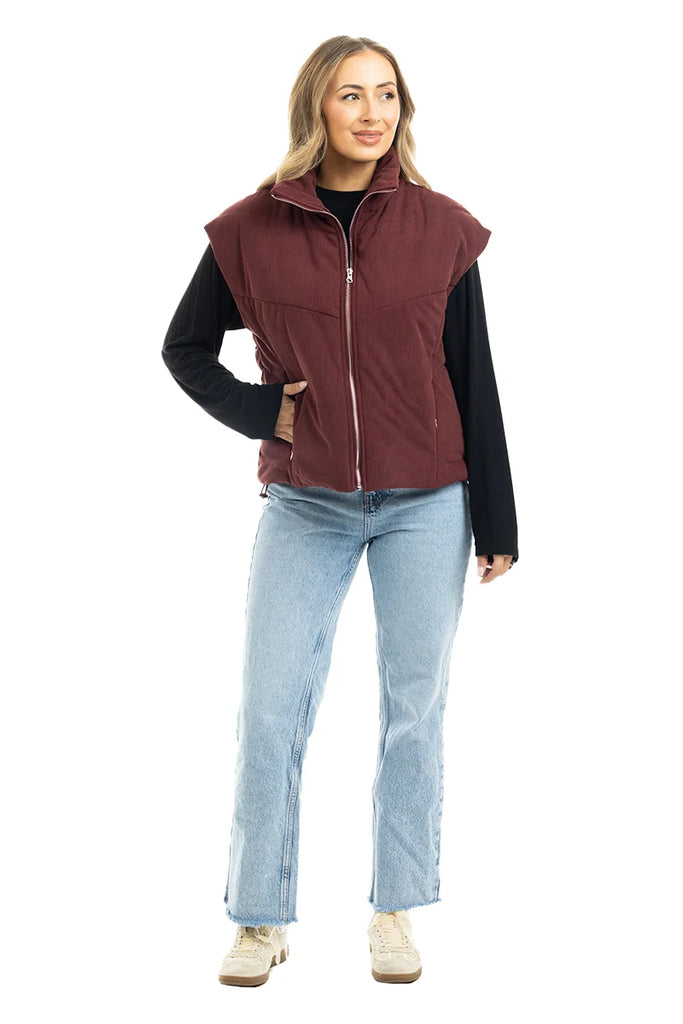 A person faces forward in a Katydid Wine Fall Corduroy Puffer Vest over a black long-sleeve shirt, paired with light blue jeans and beige sneakers. The outfit is showcased against a plain white background.