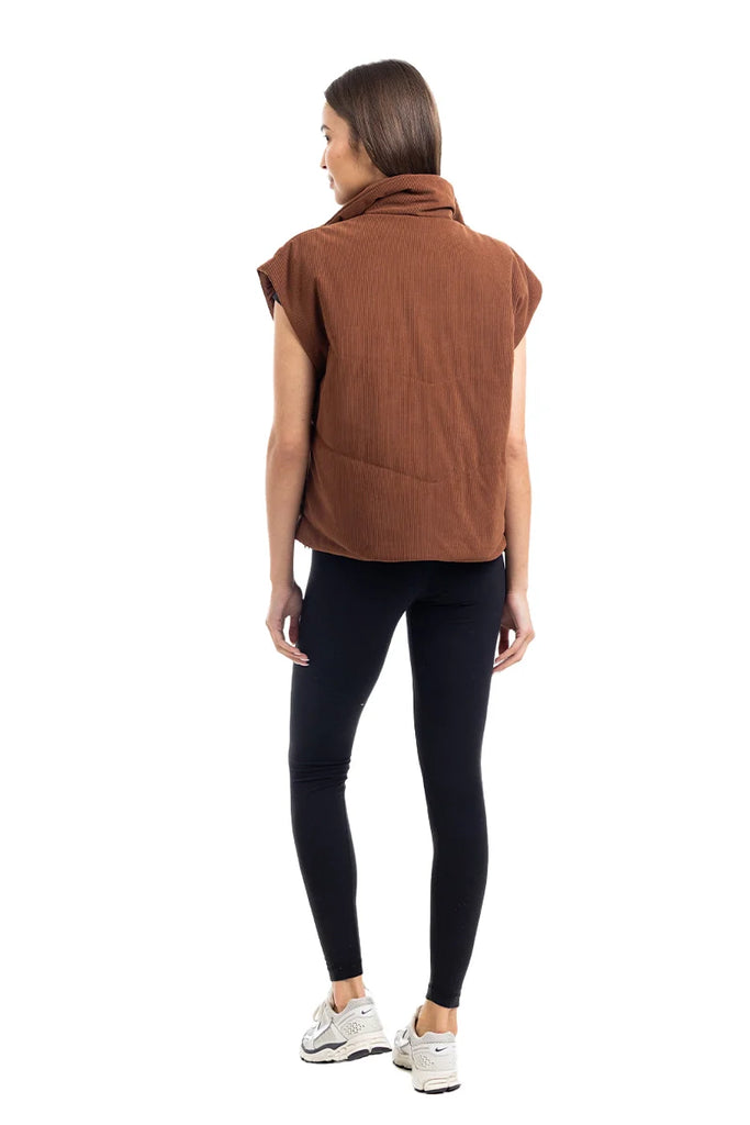 A woman faces away, wearing the Katydid Brown Corduroy Quilted Vest over a sleeveless top with black leggings and white sneakers—a stylish fall outfit set against a white background.
