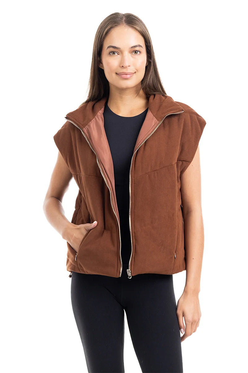 A woman with long brown hair models the Katydid Brown Corduroy Quilted Vest over a black top and leggings, posing against a white background with a slight smile—a stylish choice for fall.