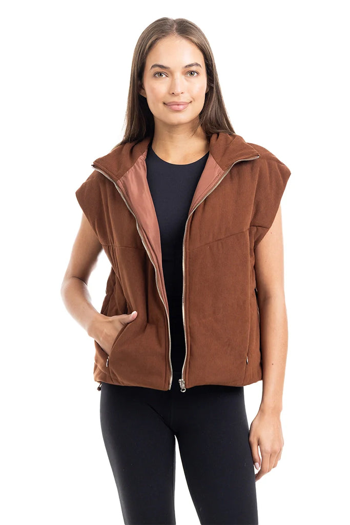 A woman with long brown hair models the Katydid Brown Corduroy Quilted Vest over a black top and leggings, posing against a white background with a slight smile—a stylish choice for fall.