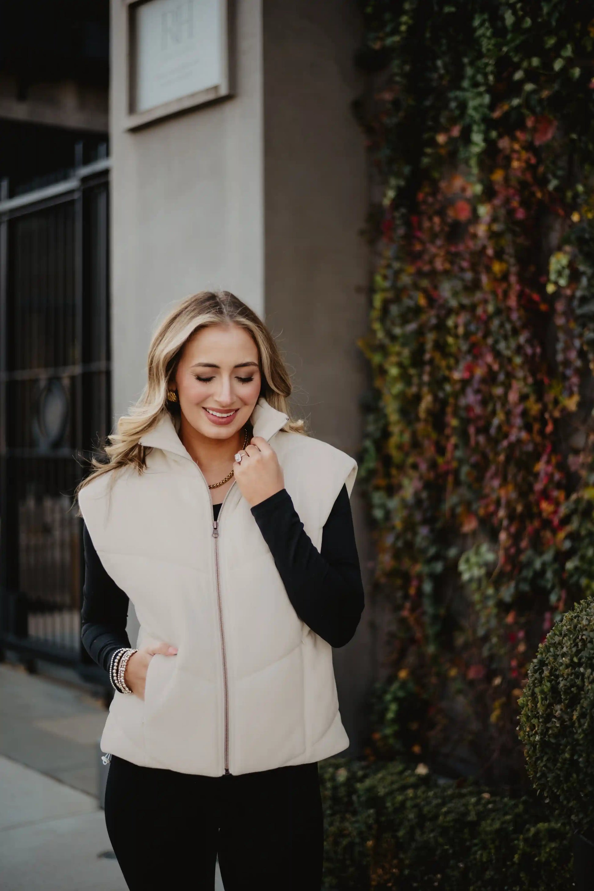 A woman with blonde hair smiles while walking outside near colorful ivy, wearing the Katydid Cloud White Corduroy Puffer Vest over a black top.