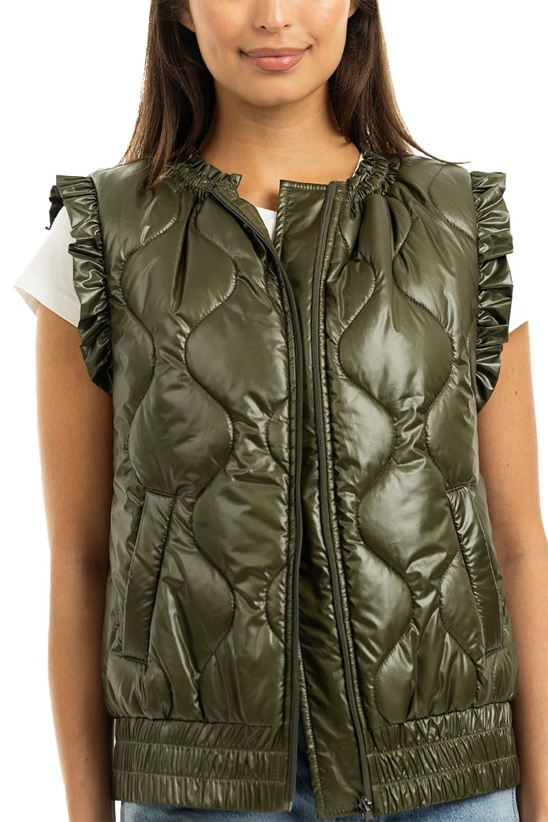 A woman wears the Katydid Olive Metallic Puffer Lightweight Vest with ruffled sleeve edges over a white short-sleeve shirt. The vest has a front zipper, side pockets, and a fitted hem. Her hair is down and she smiles slightly.