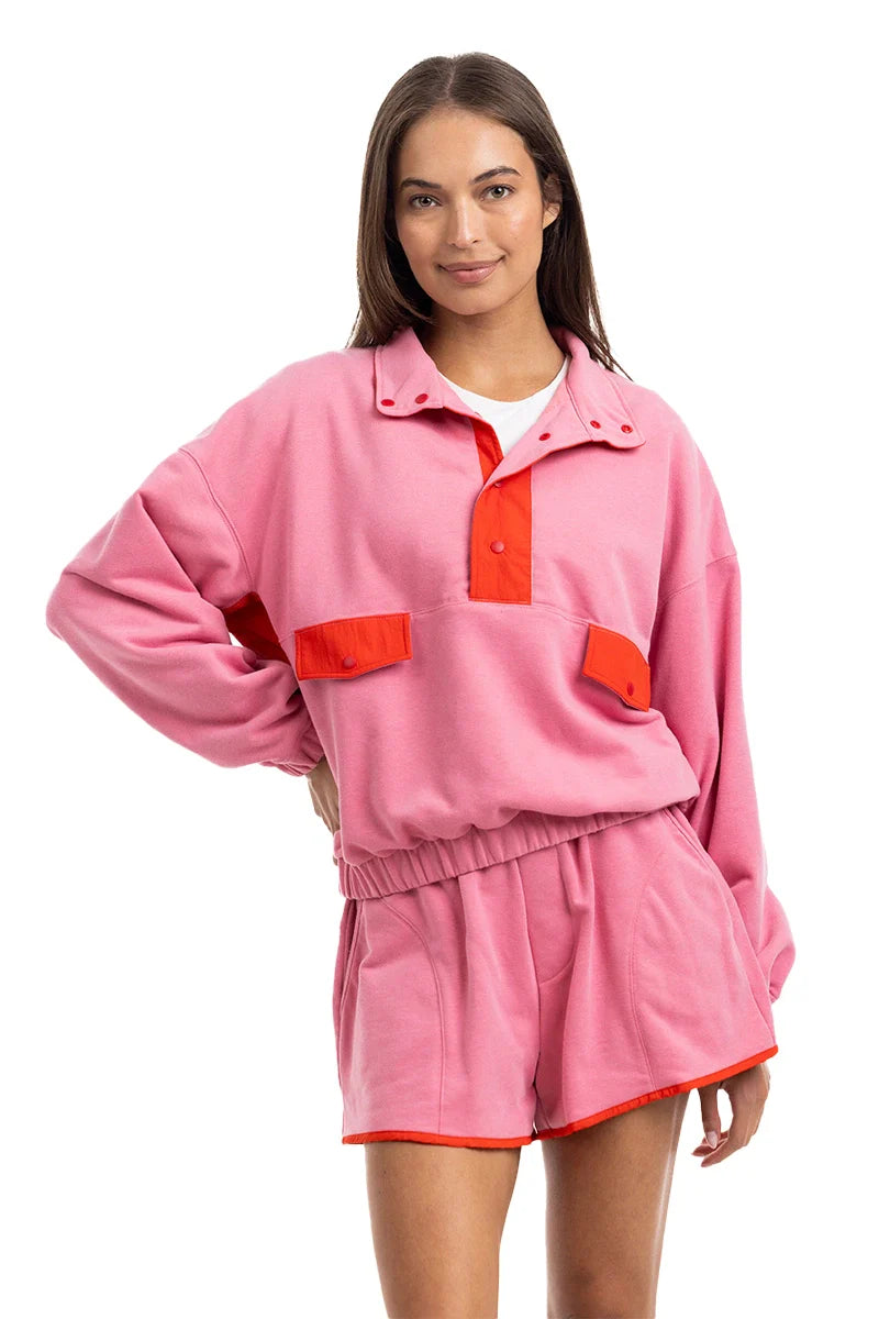 A woman smiles in the Katydid Pink  Orange Quarter Snap 2 Piece Athletic Set, featuring a loose-fit pink pullover with red accents and matching shorts made from ultra-soft performance fabric, posing confidently with one hand on her hip.