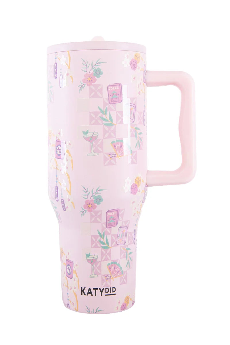 The Mahjong Pieces Hydration Tumbler by Katydid is a tall, light pink tumbler with a large handle, featuring pastel illustrations of flowers, drinks, books, and geometric shapes. The Katydid brand name is printed near the base in black letters.