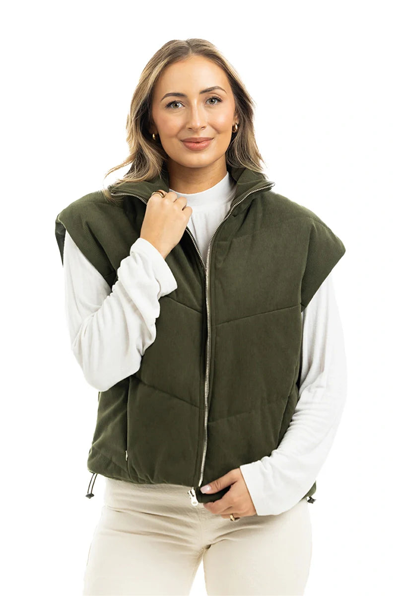 A woman with light brown hair smiles gently, standing against a white background in a Katydid Olive Corduroy Puffer Vest over a white long-sleeve shirt, with one hand touching the vest.