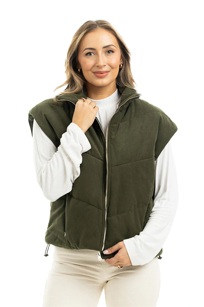 A woman with light brown hair smiles gently, standing against a white background in a Katydid Olive Corduroy Puffer Vest over a white long-sleeve shirt, with one hand touching the vest.