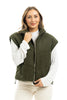 A woman with light brown hair smiles gently, standing against a white background in a Katydid Olive Corduroy Puffer Vest over a white long-sleeve shirt, with one hand touching the vest.
