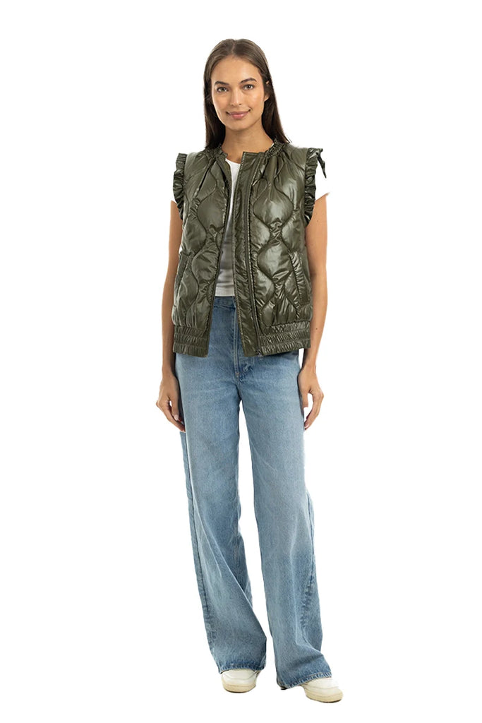 A woman faces forward in a Katydid Olive Metallic Puffer Lightweight Vest over a white top, paired with wide-leg blue jeans and white sneakers, set against a white background.
