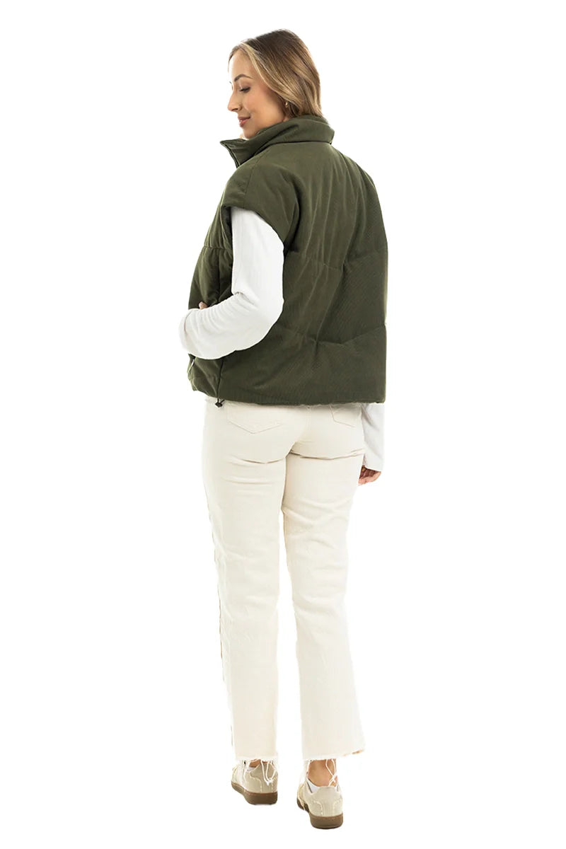 A woman models the Katydid Olive Corduroy Puffer Vest over a white long-sleeve shirt, cream pants, and beige shoes. She faces away but looks back with a smile against a white background.