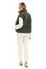 A woman models the Katydid Olive Corduroy Puffer Vest over a white long-sleeve shirt, cream pants, and beige shoes. She faces away but looks back with a smile against a white background.