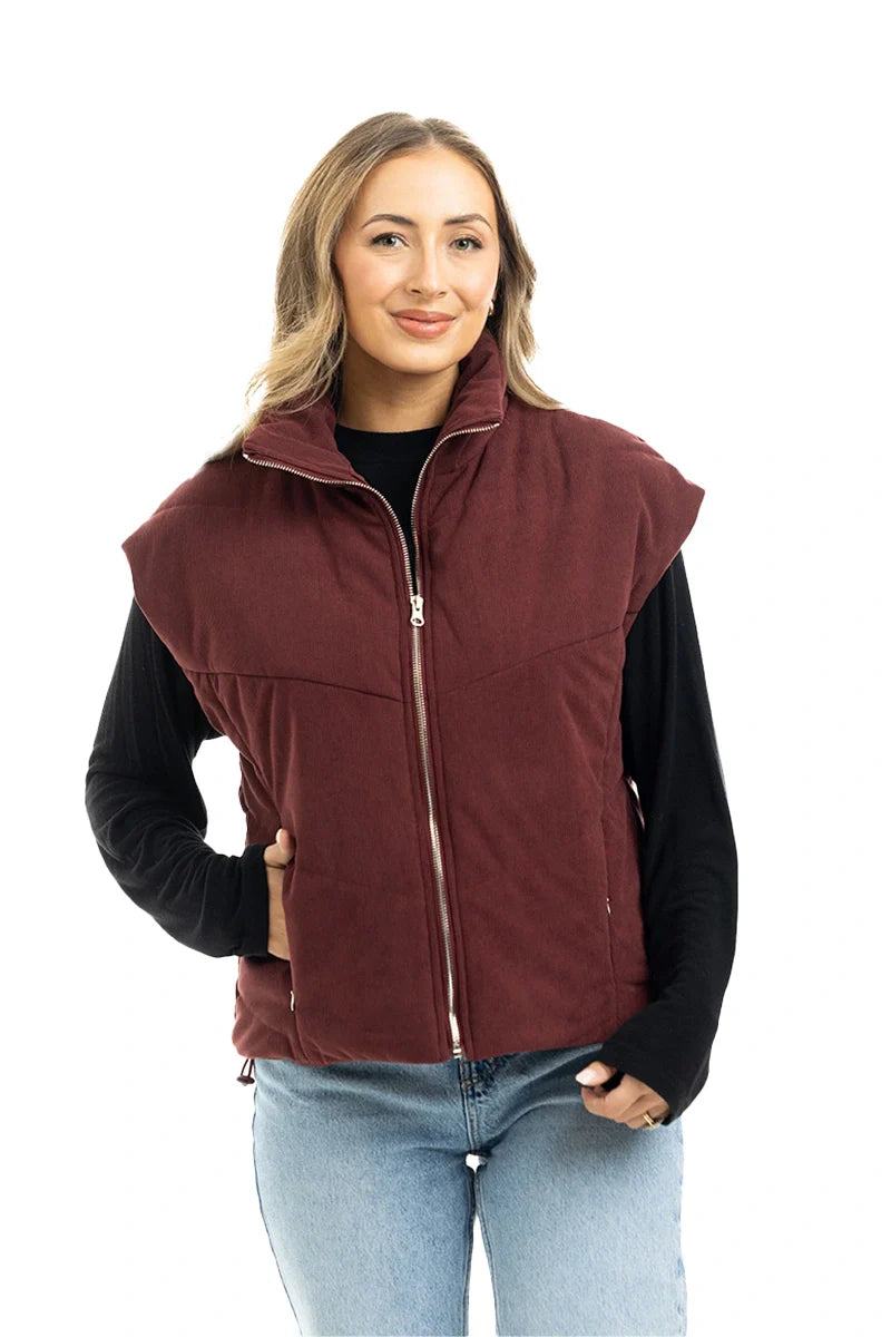 A woman smiles with one hand in her pocket, wearing the Katydid Wine Fall Corduroy Puffer Vest over a black long-sleeve shirt and light blue jeans—an ideal fall fashion look against a white background.