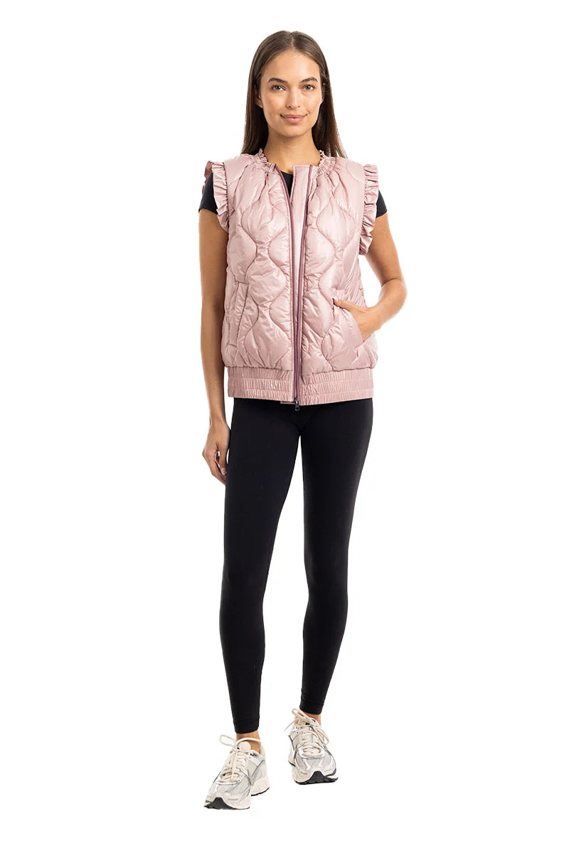 A woman faces forward against a white background, wearing the Katydid Blush Metallic Puffy Vest, black leggings, a black top, and white athletic shoes.