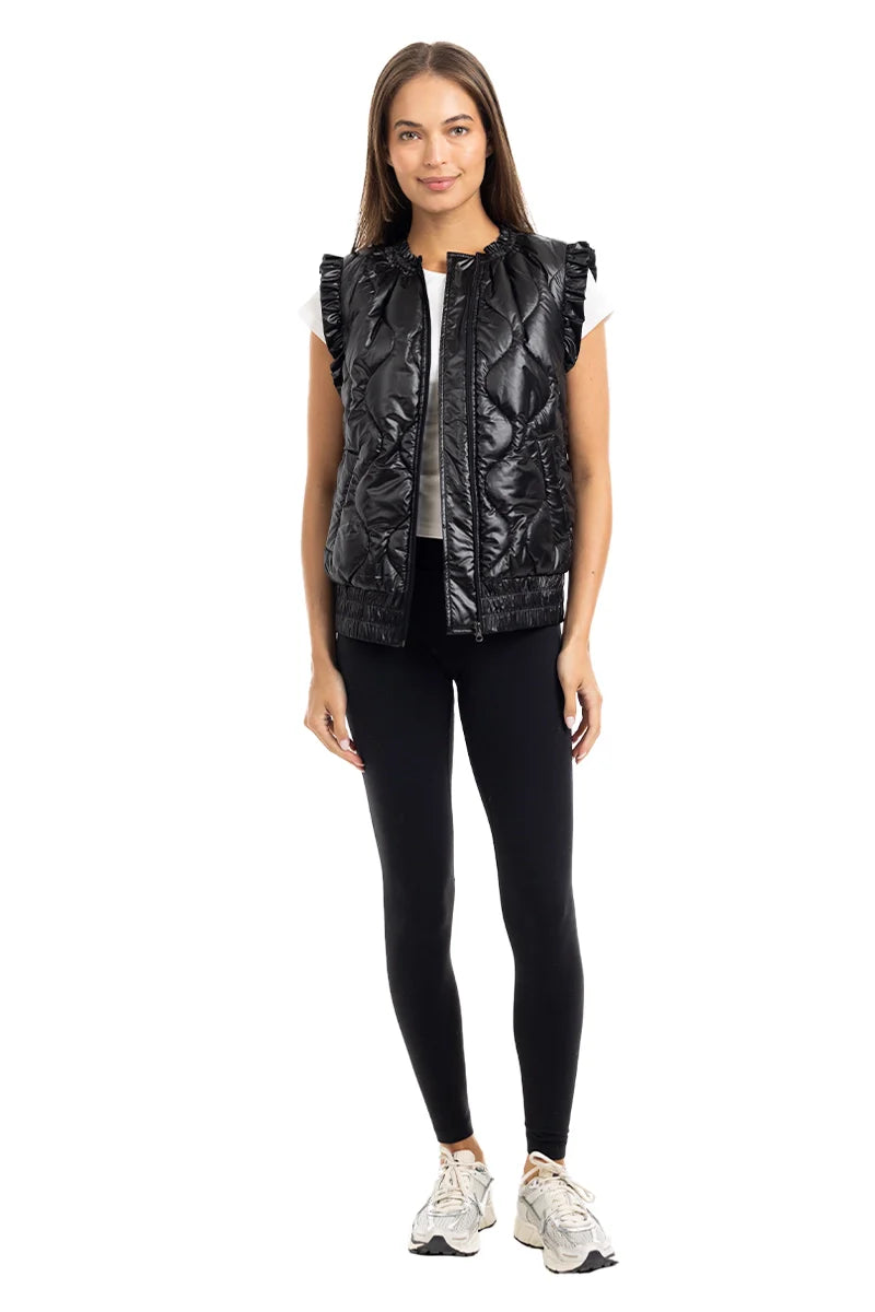A woman faces forward against a white background, wearing the Katydid Black Metallic Quilted Vest with ruffled shoulders over a white top, paired with black leggings and white sneakers—an ideal women’s fall layer.