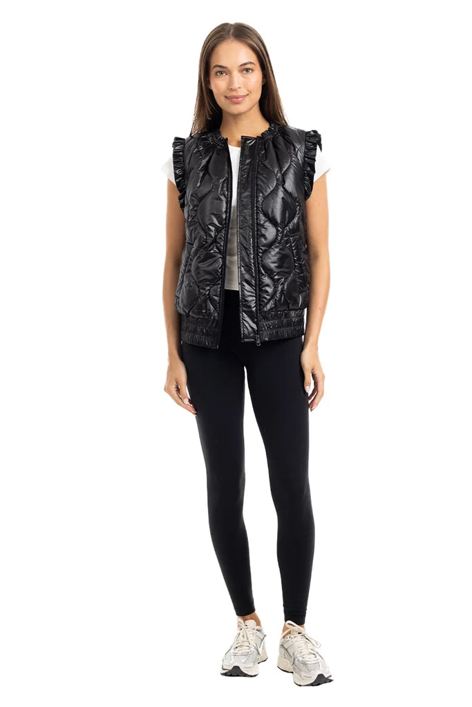 A woman faces forward against a white background, wearing the Katydid Black Metallic Quilted Vest with ruffled shoulders over a white top, paired with black leggings and white sneakers—an ideal women’s fall layer.