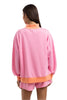 A woman with straight brown hair, seen from behind, wears the Katydid Pink  Orange Duo Mockneck Sweatset—a loose-fitting set with a pink sweatshirt featuring orange trim and matching shorts.