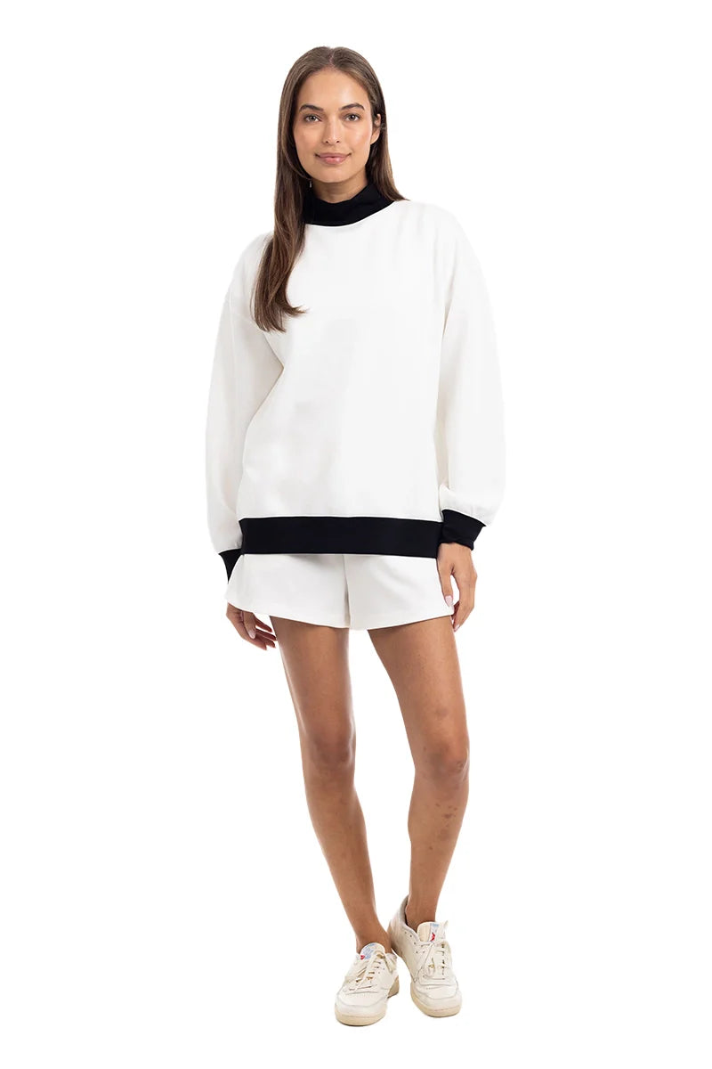 A woman stands facing forward in the Katydid White  Black Duo Mockneck Set, featuring a white long-sleeve top with black collar and hem accents, matching white shorts, and sneakers. She has long brown hair and a neutral expression against a white background.