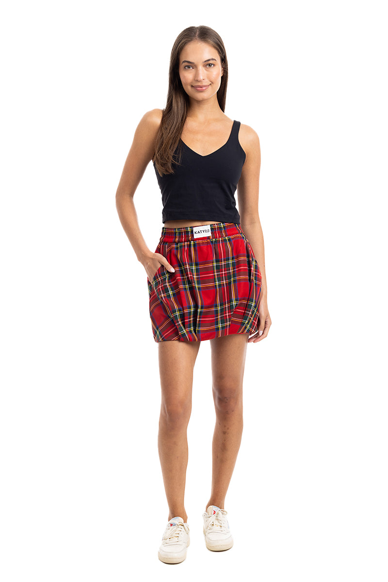 A woman stands facing forward in a sleeveless black top, Katydid Royal Stewart Plaid Mini Bubble Skirt with pockets and visible label, and white sneakers. She smiles slightly against a white background—ideal for a festive Christmas party look.