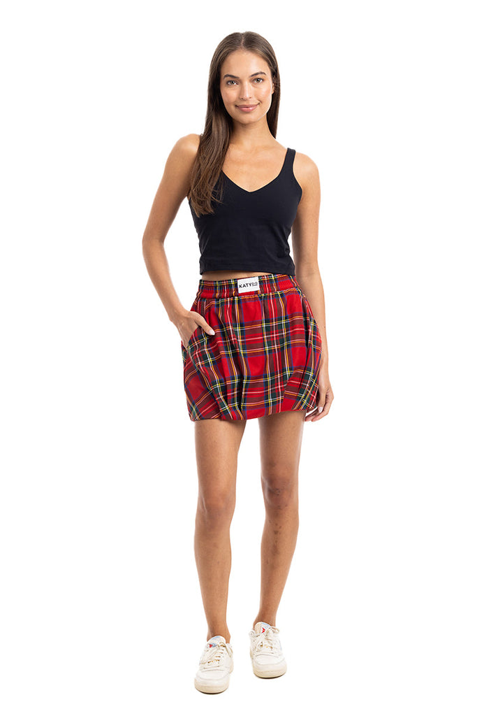 A woman stands facing forward in a sleeveless black top, Katydid Royal Stewart Plaid Mini Bubble Skirt with pockets and visible label, and white sneakers. She smiles slightly against a white background—ideal for a festive Christmas party look.