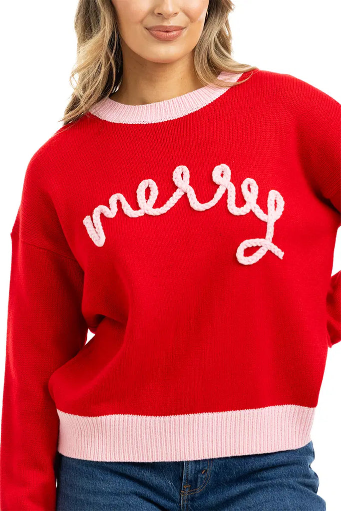 A woman with long, wavy blond hair wears the Katydid Merry Embroidered Sweater Christmas Woman—a red knit sweater with light pink trim and merry in white cursive. She is smiling slightly.