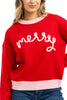 A woman with long, wavy blond hair wears the Katydid Merry Embroidered Sweater Christmas Woman—a red knit sweater with light pink trim and merry in white cursive. She is smiling slightly.